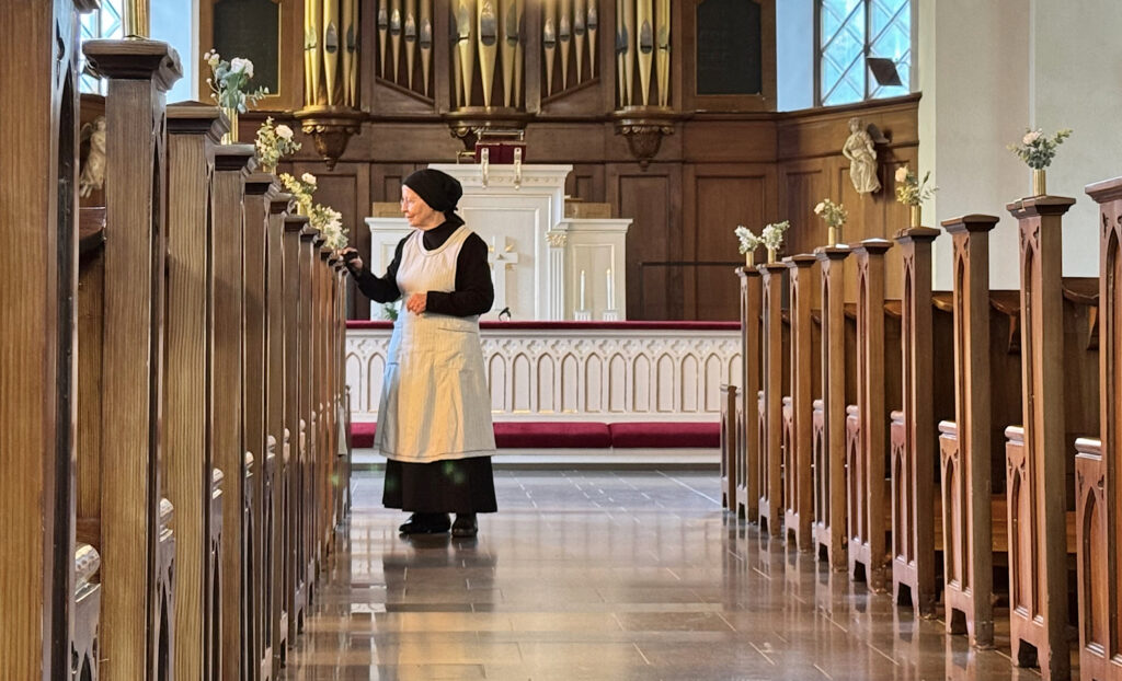 The artist, dressed in a black dress and a white apron, wiping dust of the church benches.