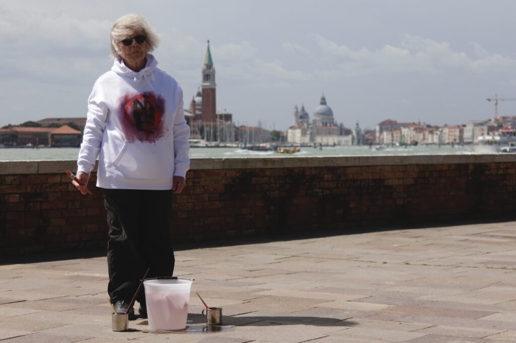 The artist stands on the bank of Giardini, Venice, Italy. She has red and black paint on her white hoodie.